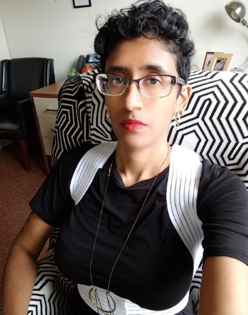 A sullen Tamil woman with short black hair and glasses sits in an office. She's wearing red lipstick and a white shoulder harness over her black shirt.