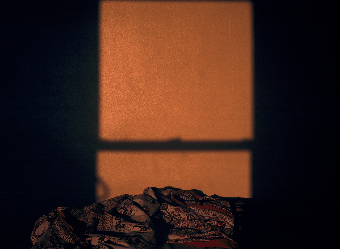 A patterned throw in a rumpled heap in a dark room, a split window of light throwing an orange wall into sharp relief between borders of shadow.