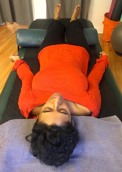 A Tamil woman in an orange long-sleeved shirt and black sweatpants, face-up on a massage table.