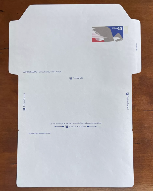 An unfolded, blank, light blue aerogram with a USA stamp, its wings labeled with numbered directions, lies on a walnut table.