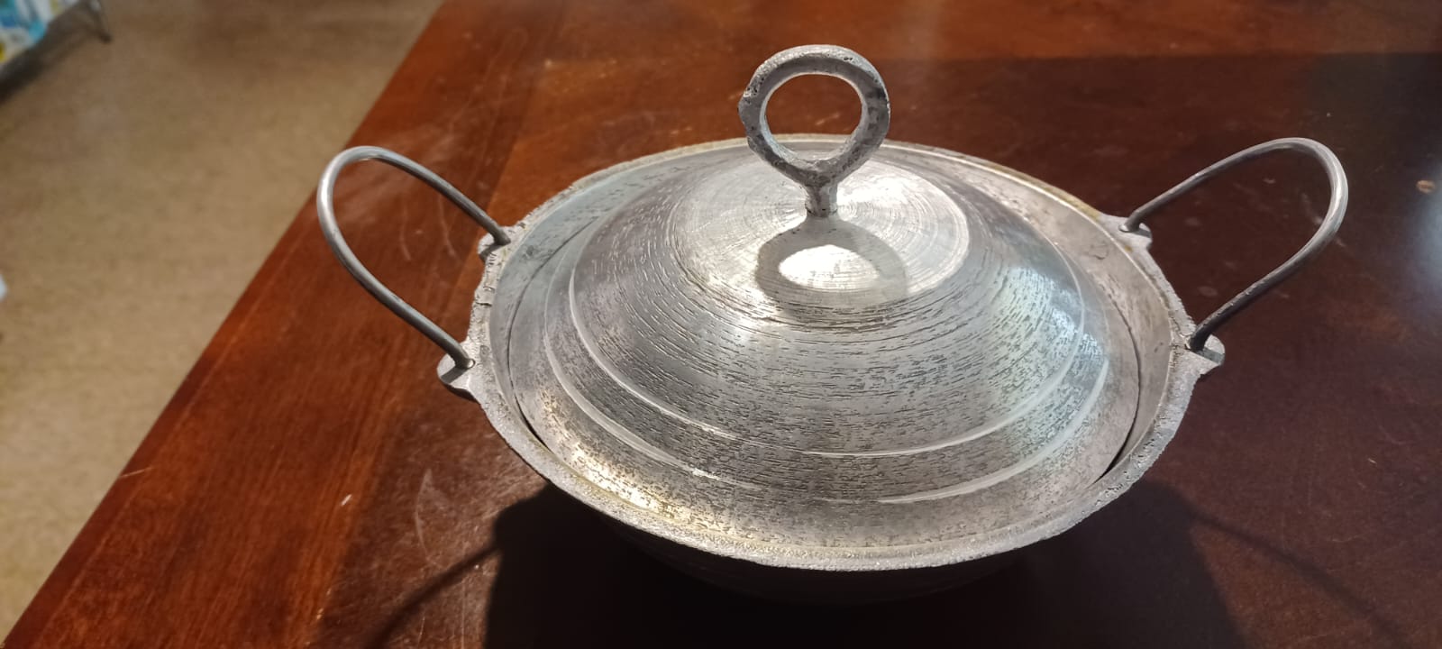 A pitted and grooved aluminum appachatti with two jutting handles and a lid with a ring handle in its center sits on a cherry veneer table.