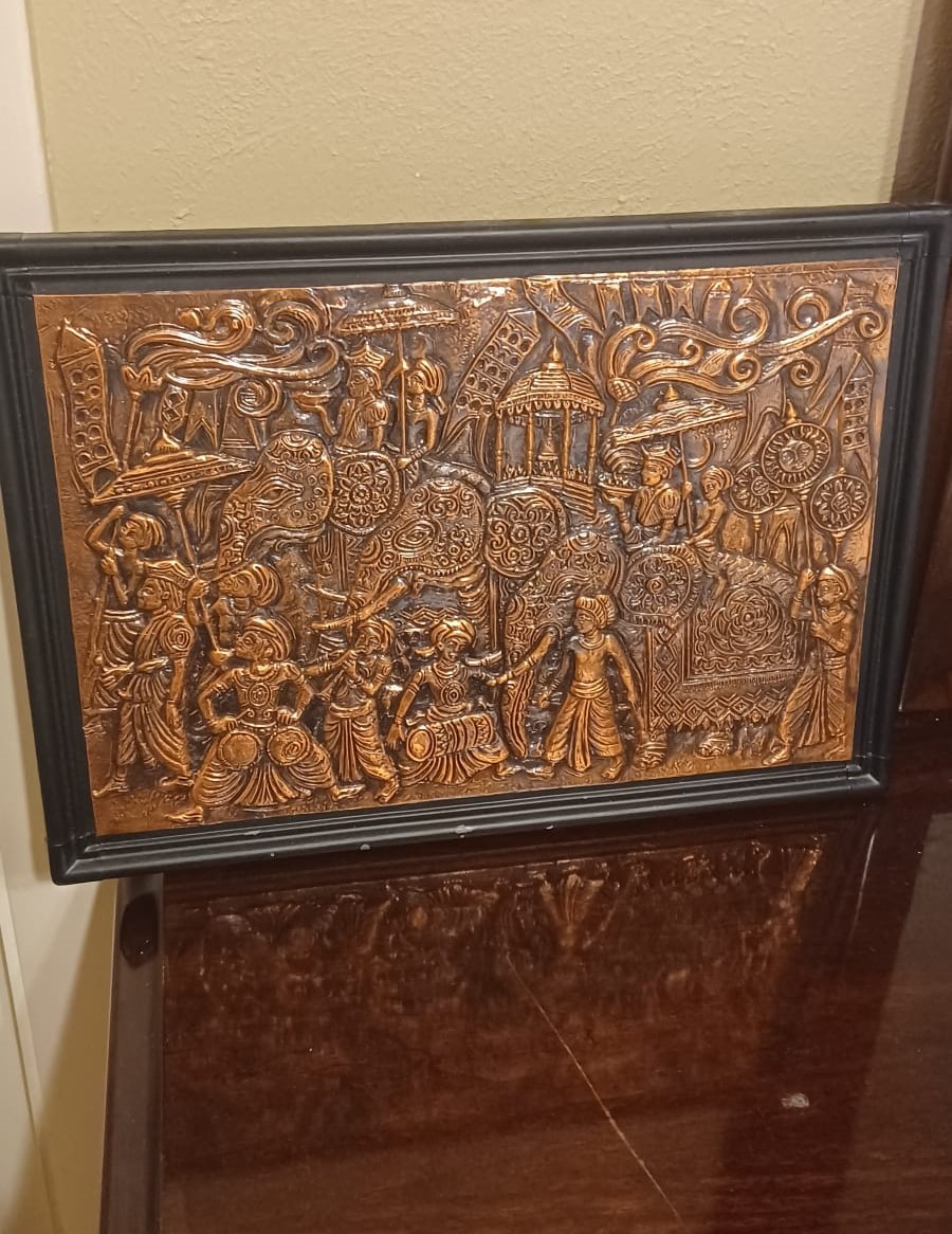 A brass repousse plaque depicts three elephants carrying royalty in canopied saddles, surrounded by drummers and footmen holding aloft banners, decorative staves, and parasols.