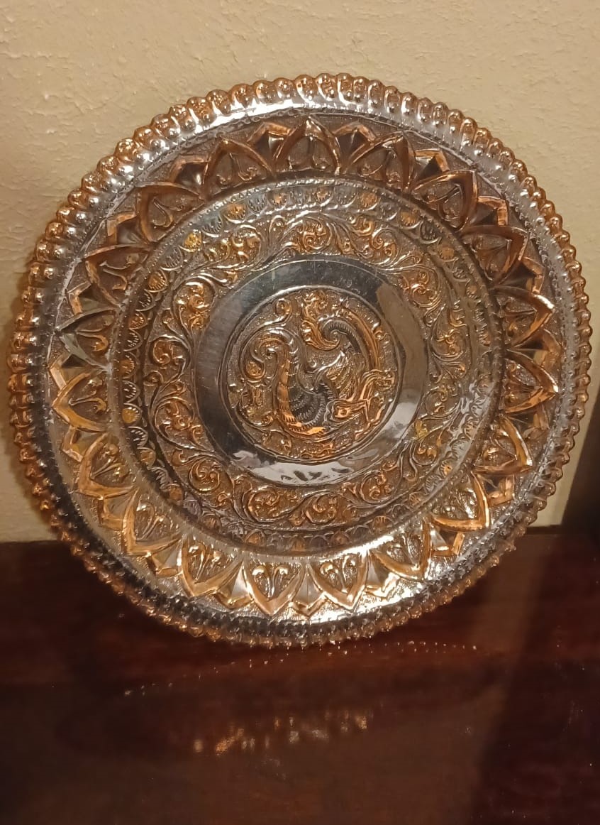A round brass plaque depicts a bird surrounded by vines and petals, with portions of the design intentionally oxidized.