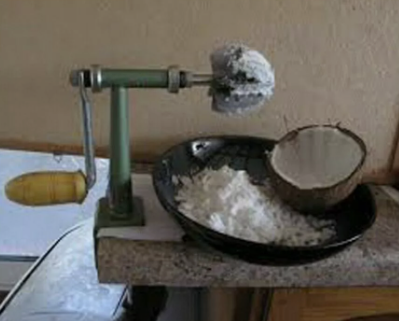A coconut scraper with a black, serrated, rotating metal blade, a wooden crank, green metal body, and vise grip is attached to a laminate counter, a black bowl full of coconut scrapings in front of it.