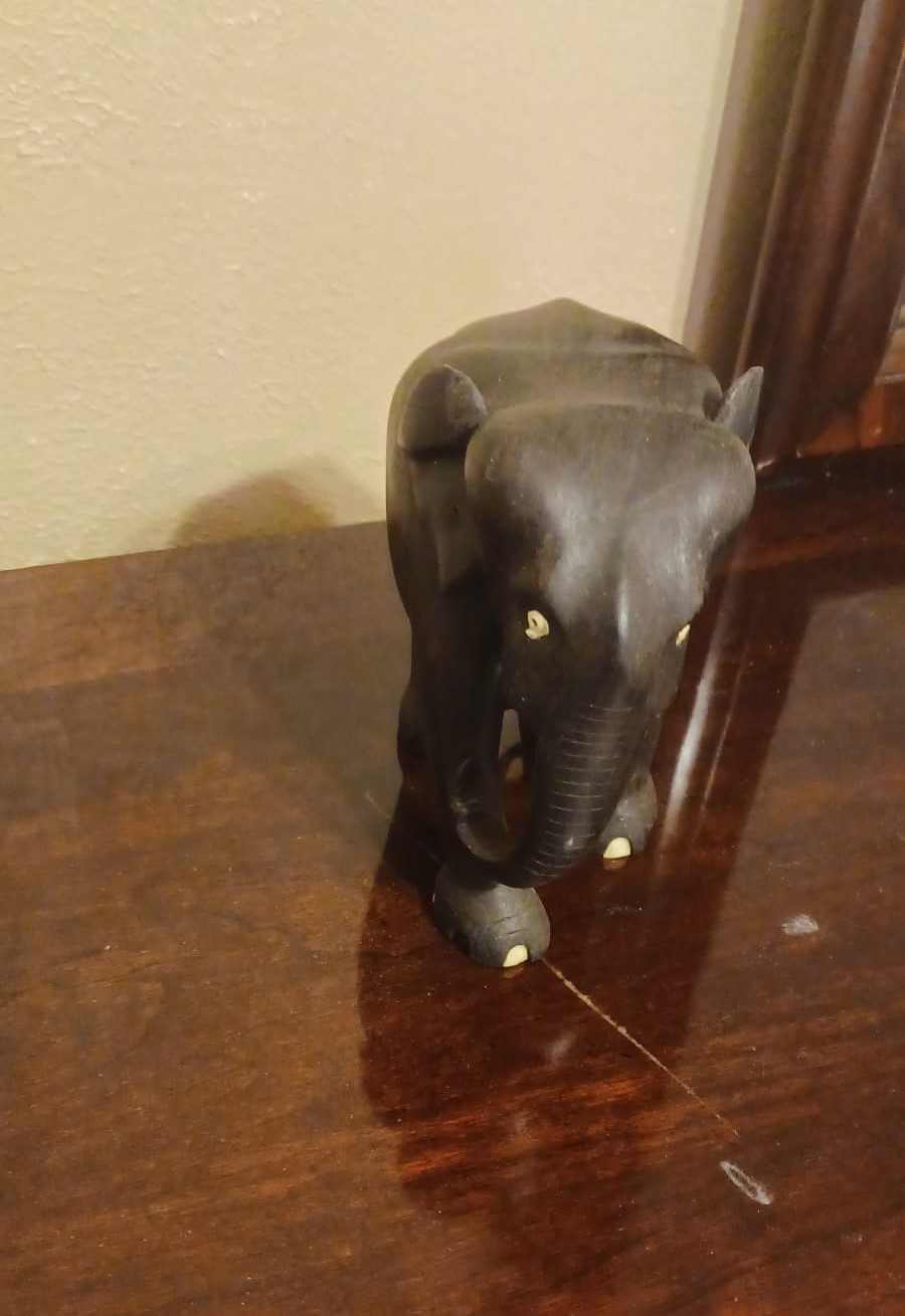 An elephant carved from ebony wood, with white eyes and nails and an inwardly curled trunk, stands on a cherry veneer table.