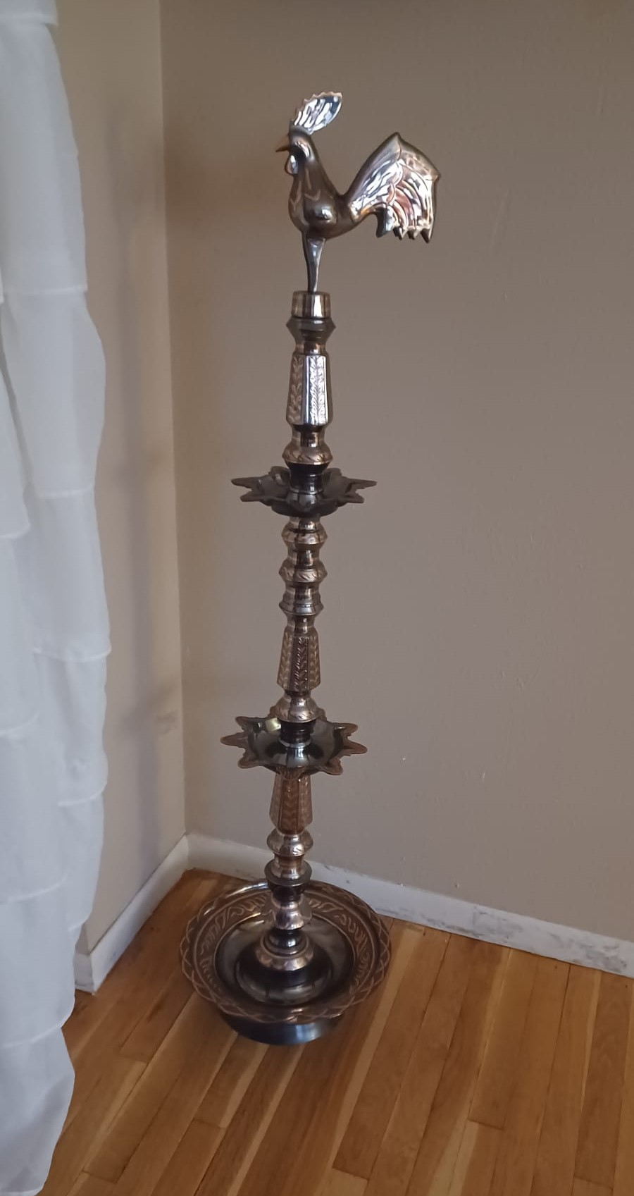 A brass two-tiered oil lamp with embossed and oxidized patterns and crowned with a Sri Lankan jungle fowl stands in an empty corner.