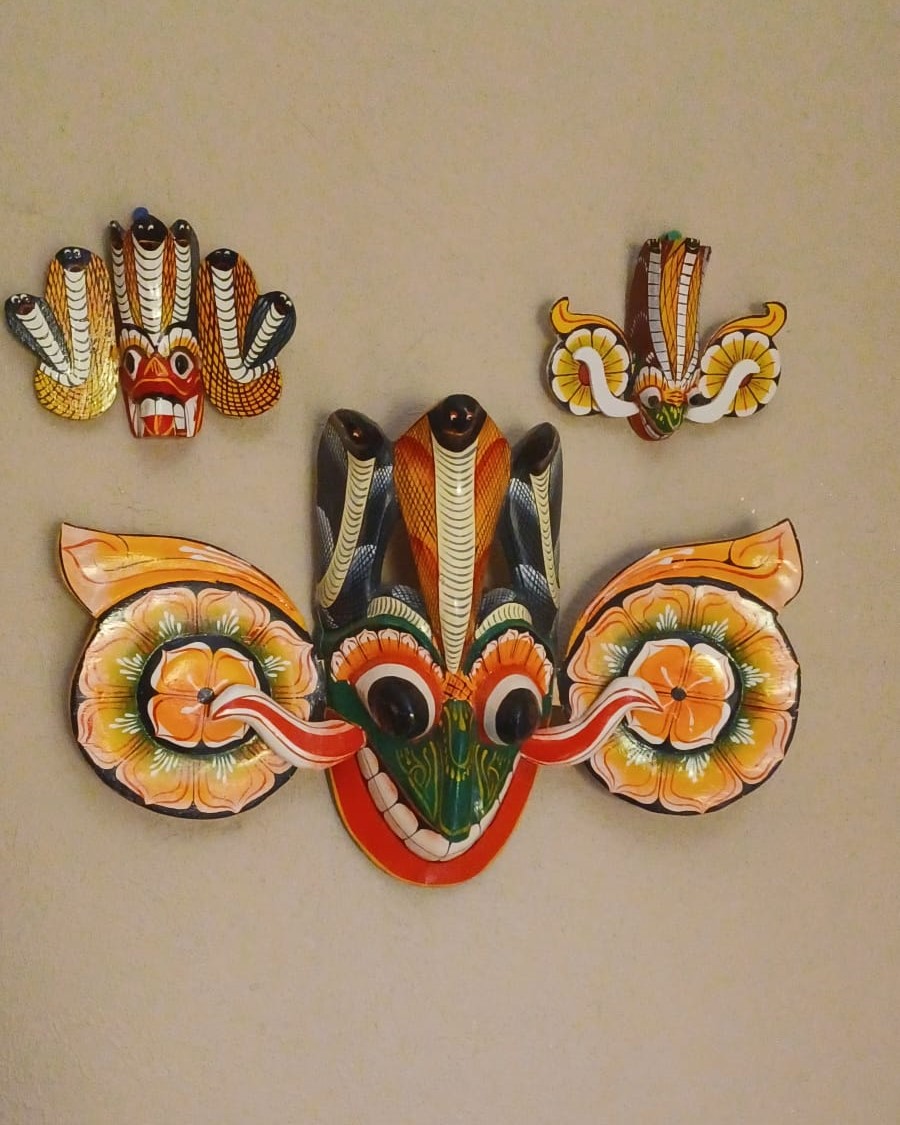 Three Sri Lankan masks, painted red, green, yellow, white, and black, with bulging eyes and protruding tongues: one small Naga Raksha mask, and one small and one large Gara Raksha mask.