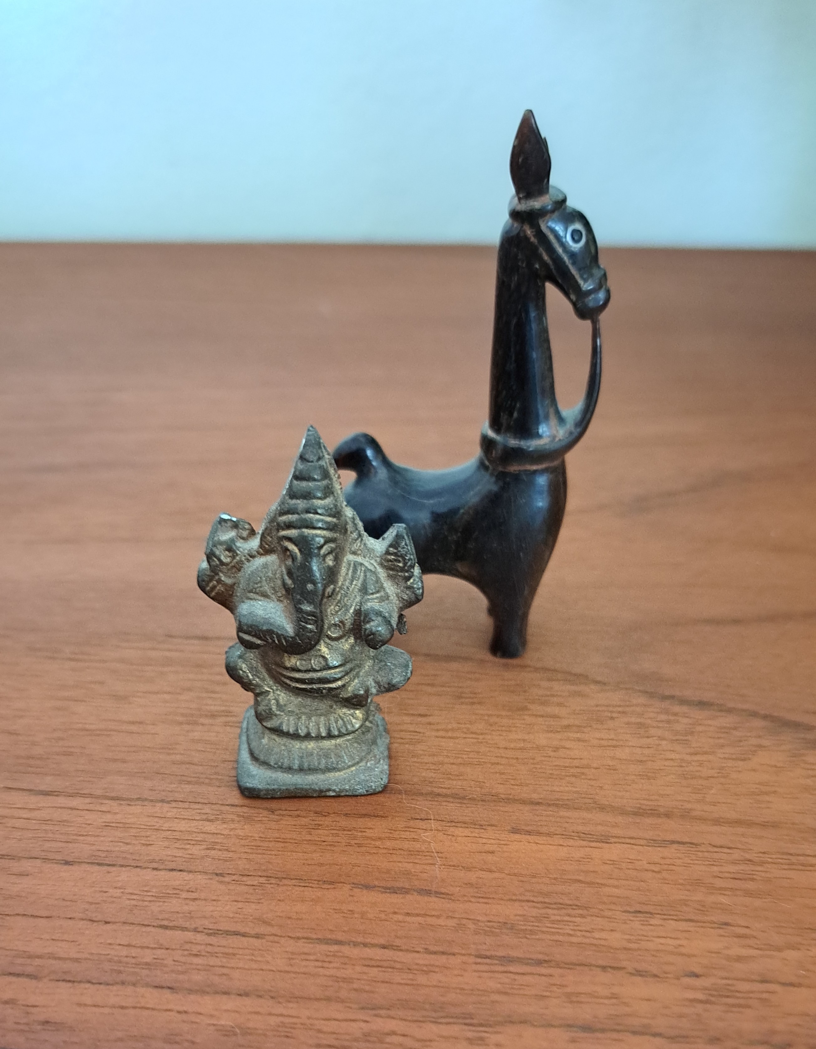 A small oxidized brass statuette of Pillaiyar and an ebony wood carving of a horse with perked up ears, a white eye, and reins draped over its neck.