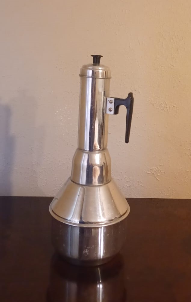 An aluminum puttu kulal, with a tall, narrow, cylindrical top, a wide base, a black plastic handle, and a lid with a black knob sits on a cherry veneer table.