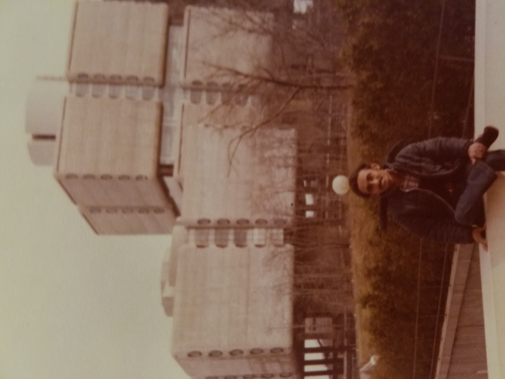 A Tamil man wit black hair and a mustache poses on a low wall in a dark blue windbreaker, a plaid shirt, and jeans in front of Stony Brook University Hospital.