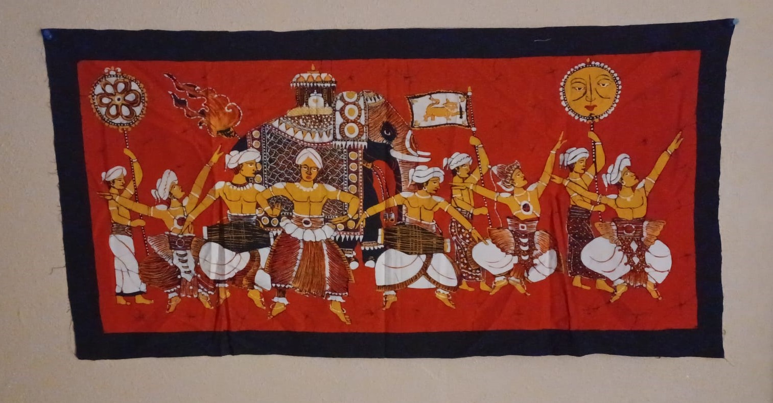 A batik tapestry with a thick, fraying black border and a red background, depicting a Kandyan perahera procession with dancers and flag- and staff-bearers surrounding a decorated elephant bearing a canopied carriage.